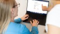 Foto von zwei Personen vor einem Laptop, wo eine Person auf das Display deutet, auf dem Grafiken zu erkennen sind.